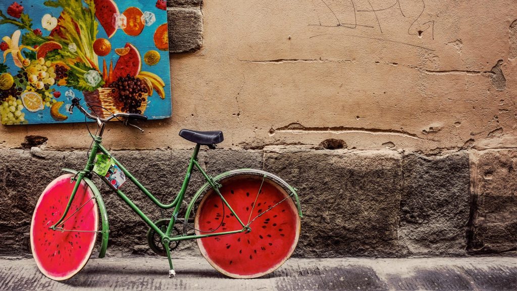 Green bicycle with watermelon-slice wheels parked by a textured wall beneath a colorful fruit-themed mural.